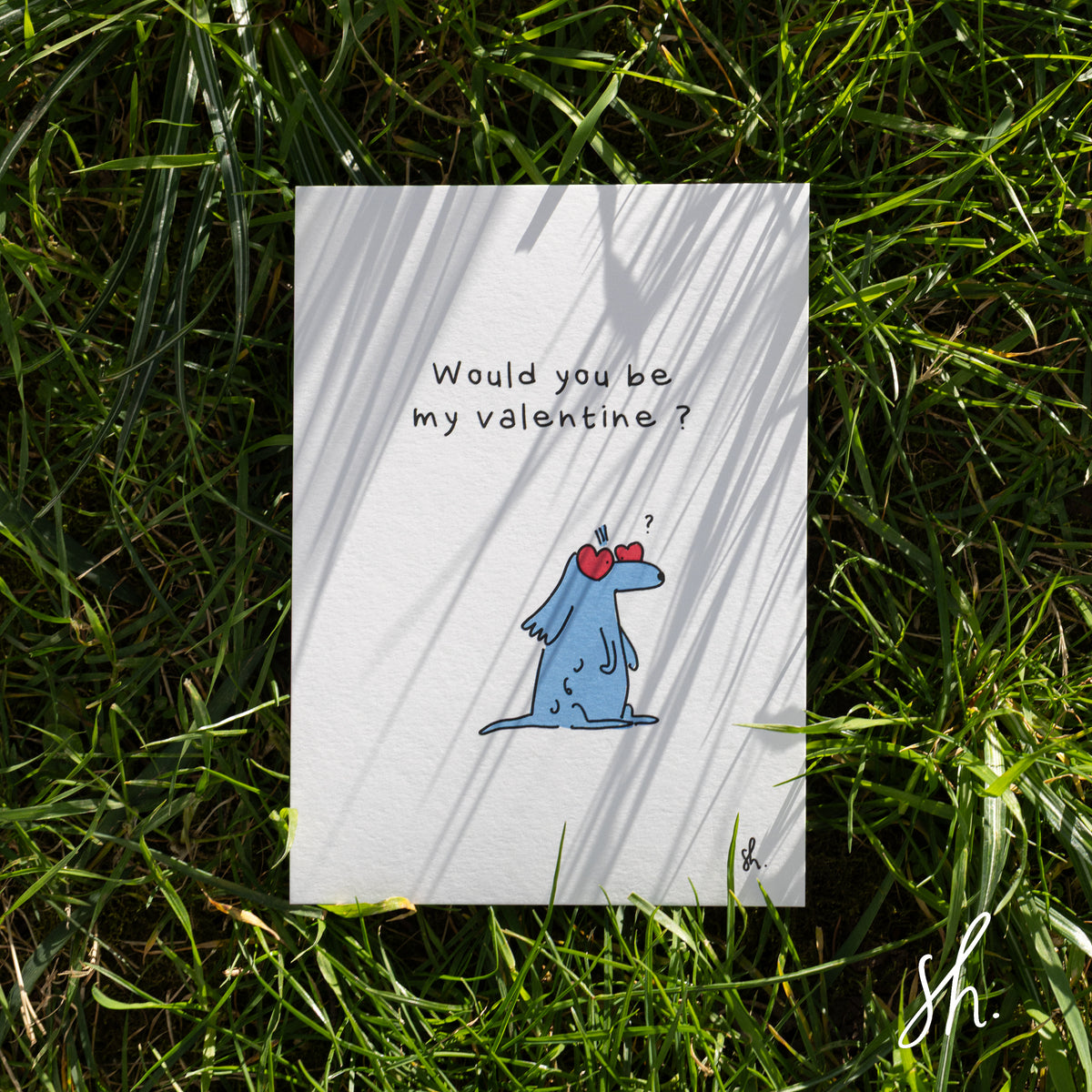 carte postale posée dans l'herbe sous un joli soleil qui reflète l'ombre de l'herbe dessus, un cocker est dessiné dessus qui porte des lunettes en forme de coeur et demande "would you be my valentine?"