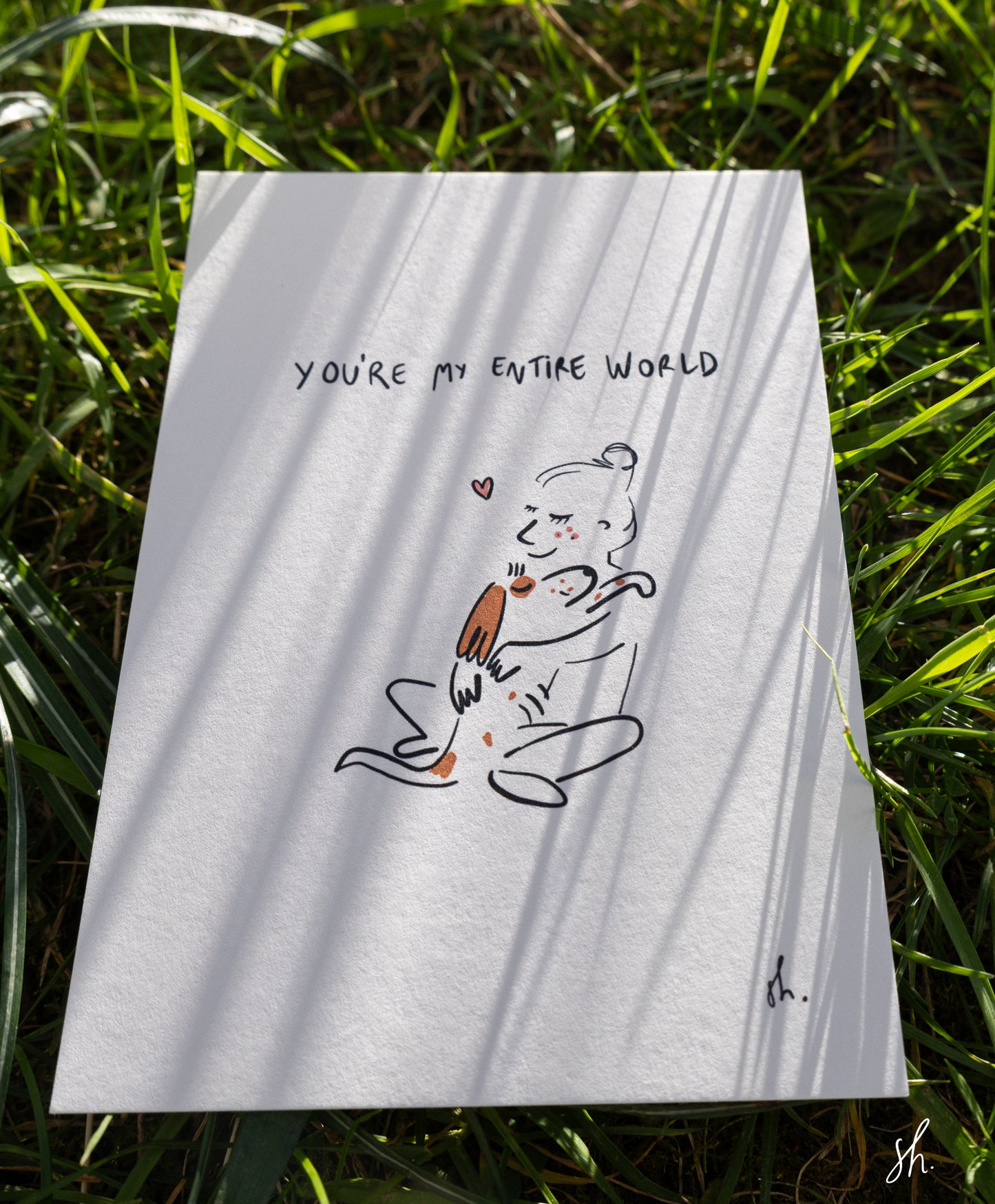 carte postale posée dans l'herbe sous un joli soleil qui reflète l'ombre de l'herbe dessus, il est dessiné un cocker qui fait un câlin à sa maitresse et est ecrit au dessu "you're my entire world" 