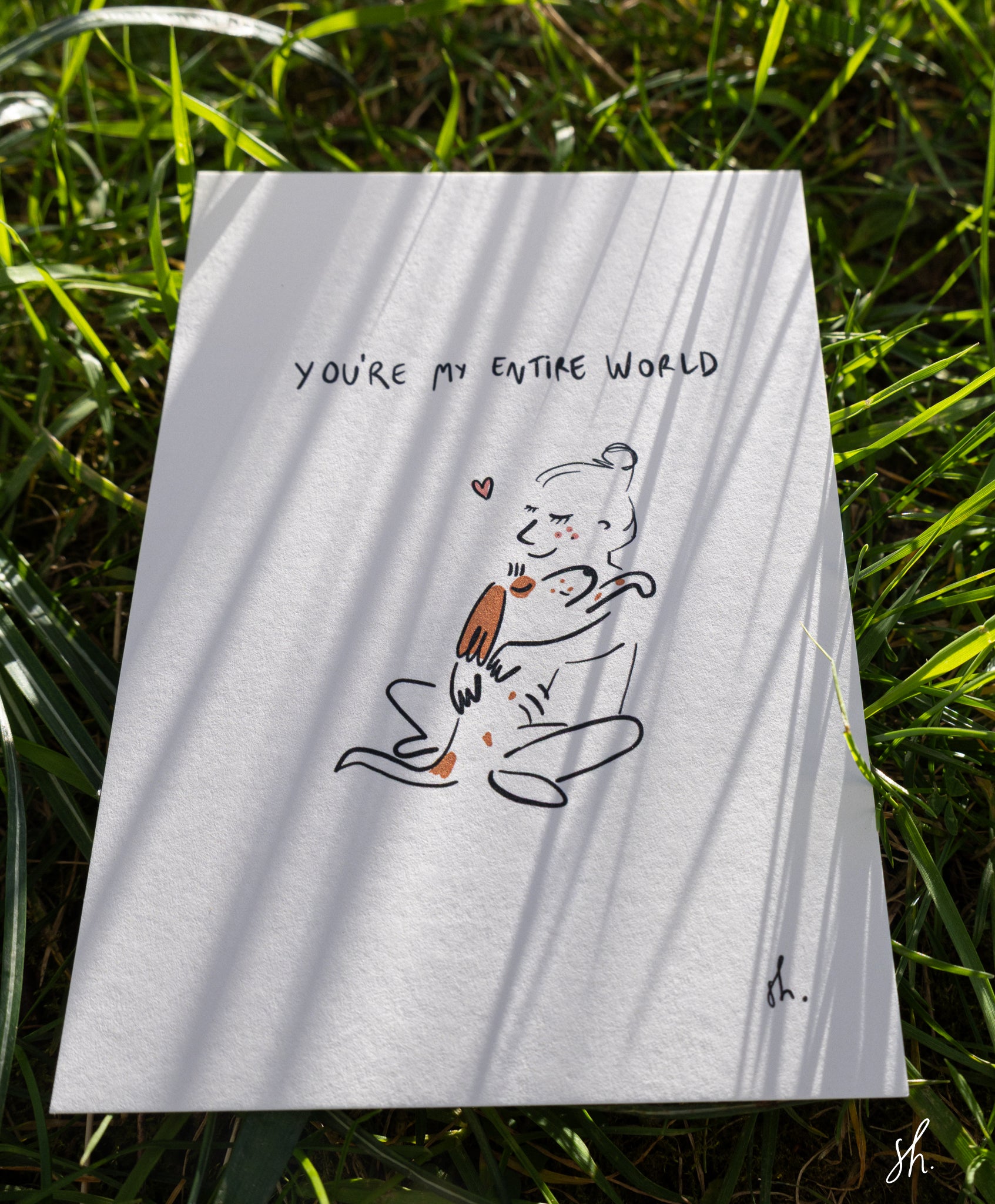 carte postale posée dans l'herbe sous un joli soleil qui reflète l'ombre de l'herbe dessus, il est dessiné un cocker qui fait un câlin à sa maitresse et est ecrit au dessu "you're my entire world" 
