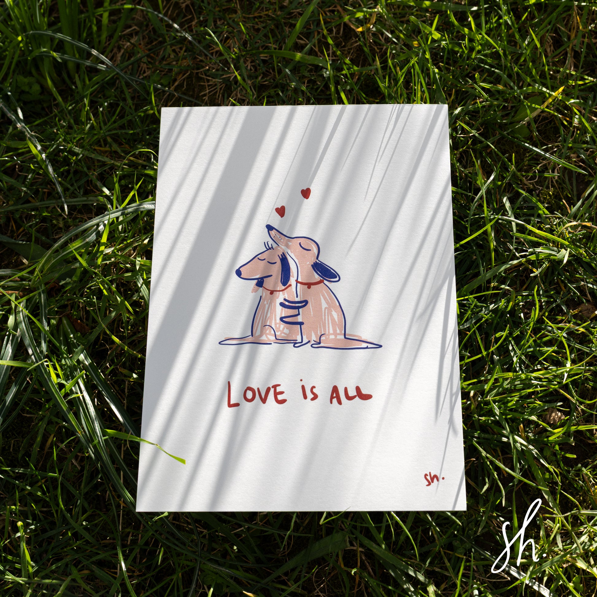 carte postale prise en photo dans l'herbe sur laquelle se reflète les ombres de l'herbe autour, deux chiens se font un câlin dessiné en rose et il est écrit en dessous 'love is all"