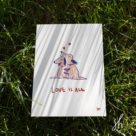 carte postale prise en photo dans l'herbe sur laquelle se reflète les ombres de l'herbe autour, deux chiens se font un câlin dessiné en rose et il est écrit en dessous 'love is all"