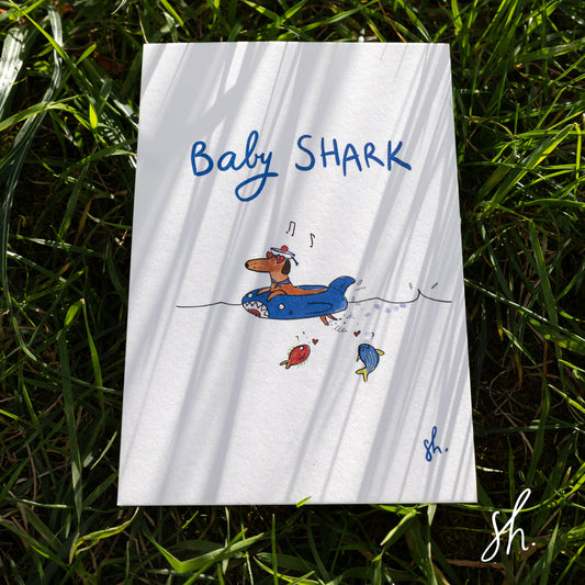petite carte posale posé dans l'herbe sous un rayon de soleil, l'herbe dessine de jolies ombres sur la carte qui représente un teckel qui nage dans une bouée en chantant baby shark avec un petit bonnet de marin et des poissons en dessous 
