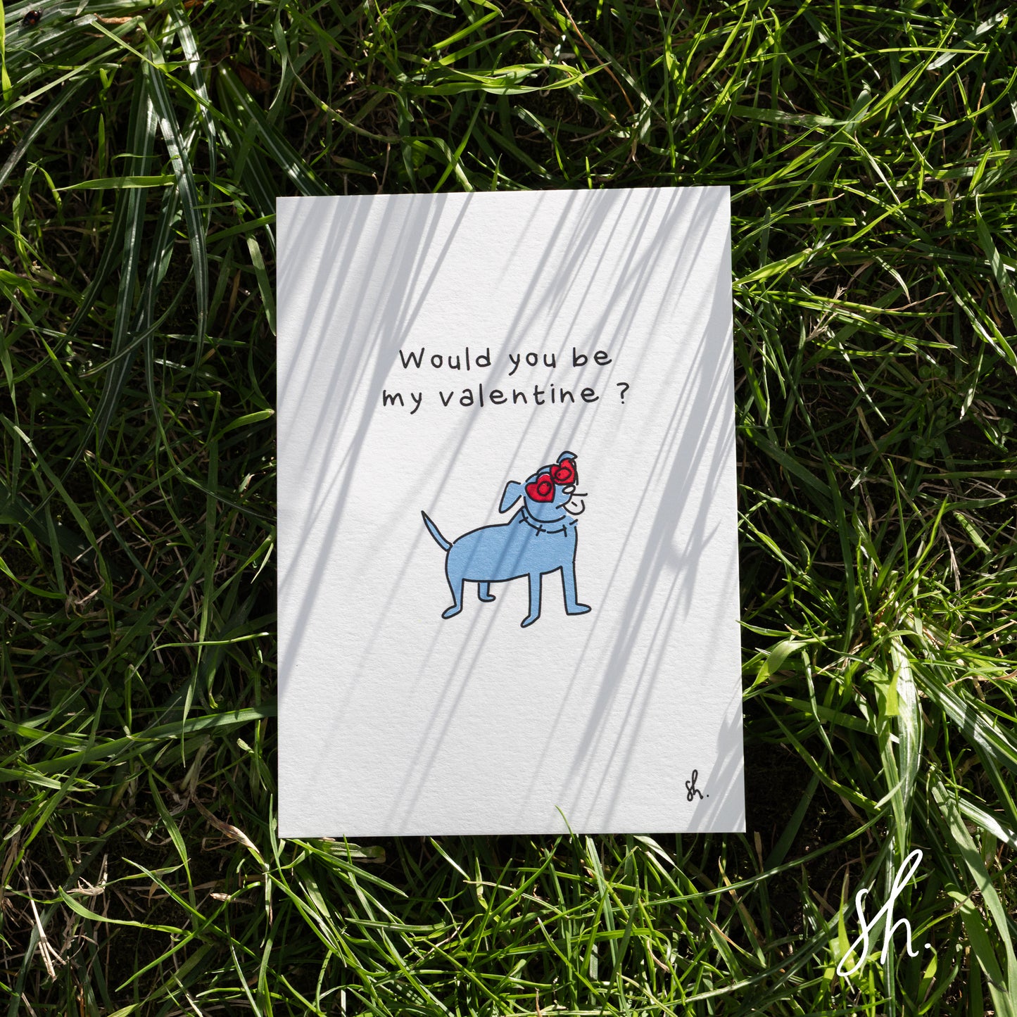 carte postale posée dans l'herbe sous un joli soleil qui reflète l'ombre de l'herbe dessus, un staffy est dessiné dessus qui porte des lunettes en forme de coeur et demande "would you be my valentine?" 