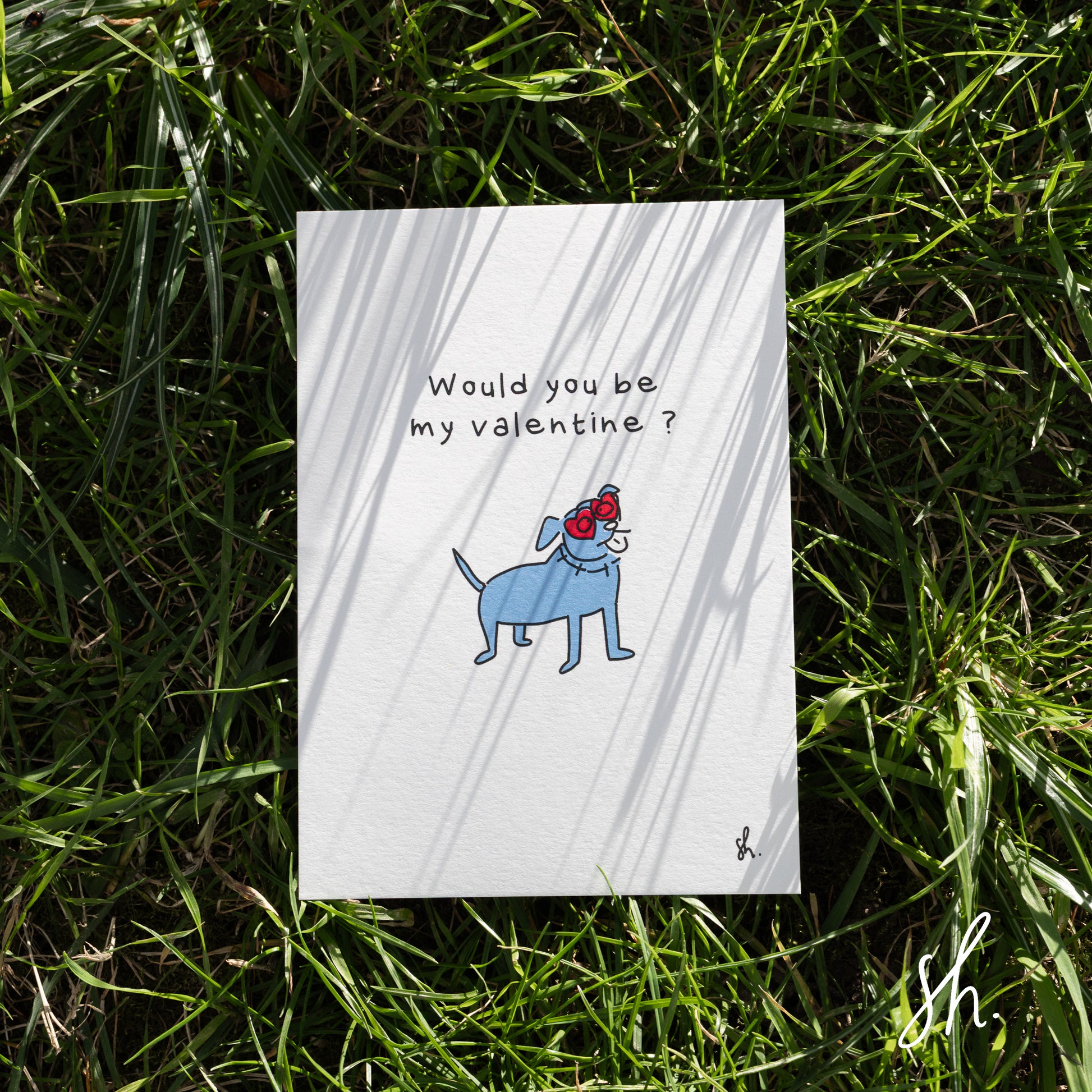 carte postale posée dans l'herbe sous un joli soleil qui reflète l'ombre de l'herbe dessus, un staffy est dessiné dessus qui porte des lunettes en forme de coeur et demande "would you be my valentine?" 