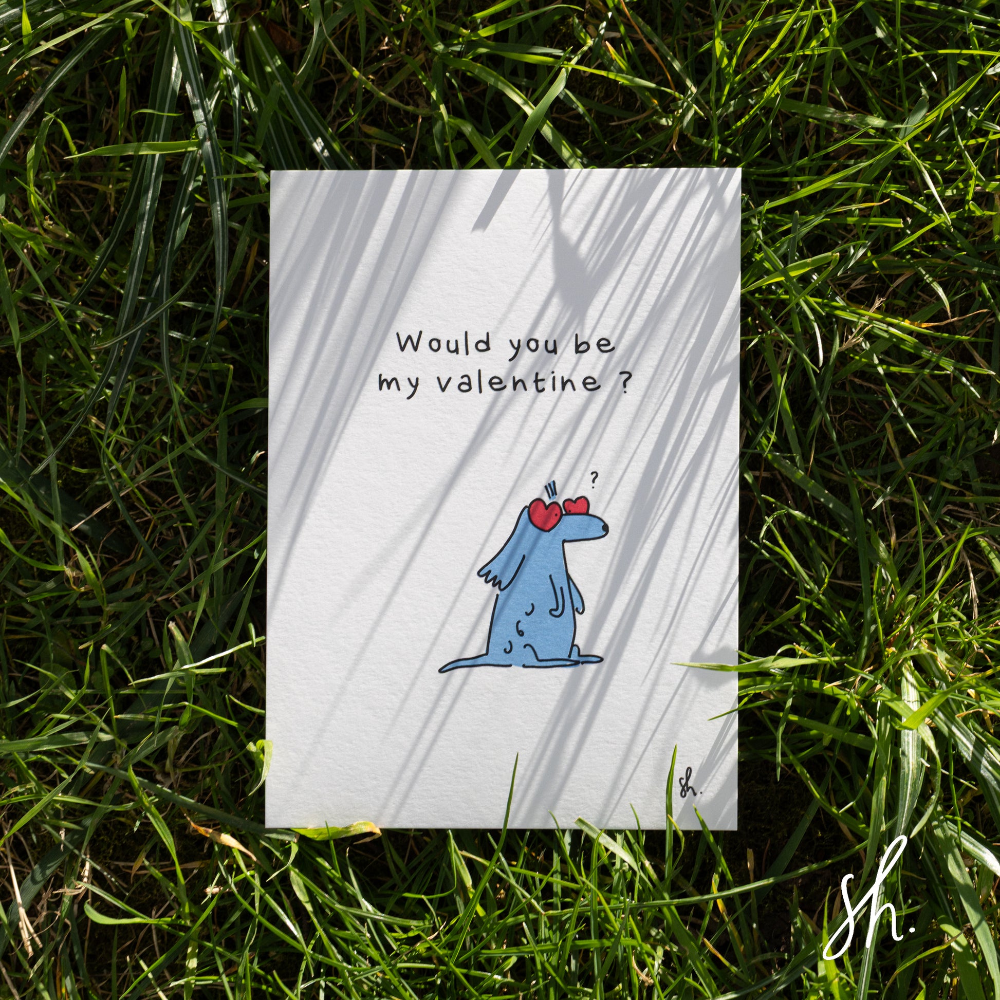 carte postale posée dans l'herbe sous un joli soleil qui reflète l'ombre de l'herbe dessus, un cocker est dessiné dessus qui porte des lunettes en forme de coeur et demande "would you be my valentine?" 