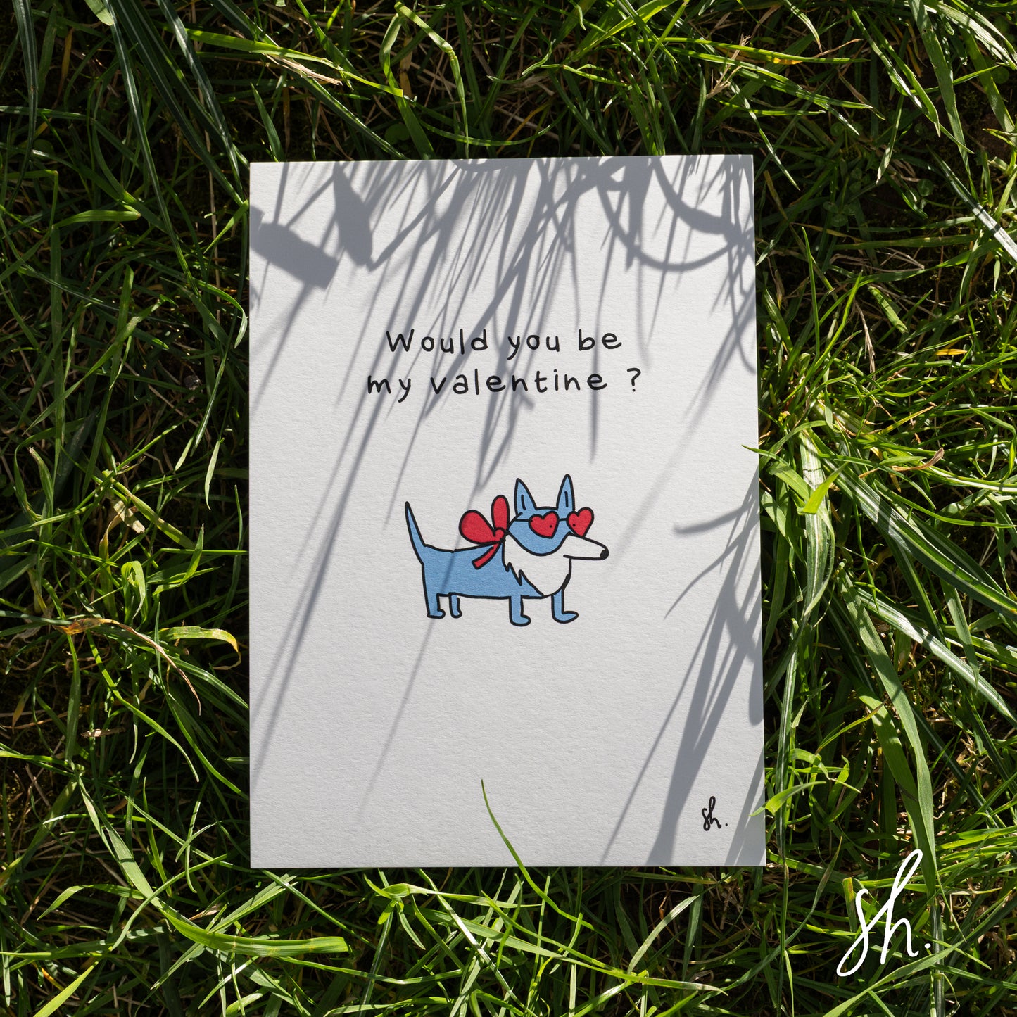 carte postale posée dans l'herbe sous un joli soleil qui reflète l'ombre de l'herbe dessus, un corgy est dessiné dessus qui porte un petit noeud rouge et des lunettes en forme de coeur et demande "would you be my valentine?" 