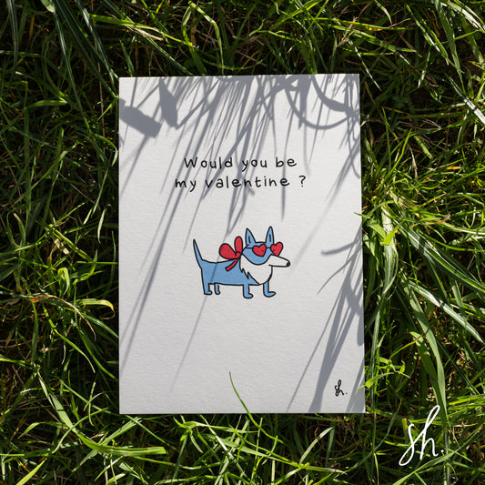carte postale posée dans l'herbe sous un joli soleil qui reflète l'ombre de l'herbe dessus, un corgy est dessiné dessus qui porte un petit noeud rouge et des lunettes en forme de coeur et demande "would you be my valentine?" 