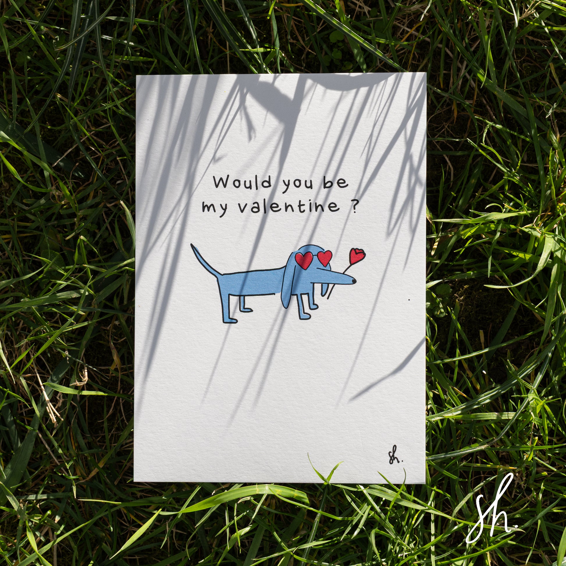 carte postale posée dans l'herbe sous un joli soleil qui reflète l'ombre de l'herbe dessus, un teckel est dessiné dessus qui porte des lunettes en forme de coeur et une rose à la bouche et demande "would you be my valentine?" 