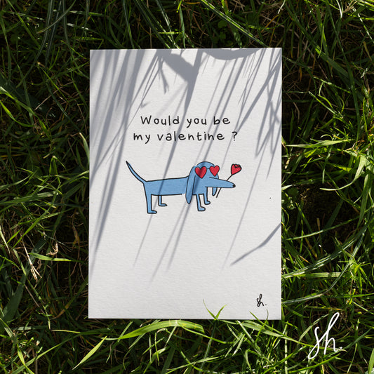 carte postale posée dans l'herbe sous un joli soleil qui reflète l'ombre de l'herbe dessus, un teckel est dessiné dessus qui porte des lunettes en forme de coeur et une rose à la bouche et demande "would you be my valentine?" 